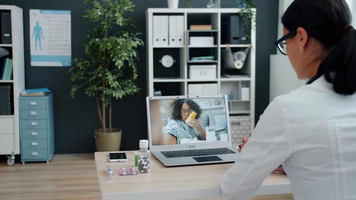 A clinician in a white coat speaking with a patient by video call during a telehealth consultation