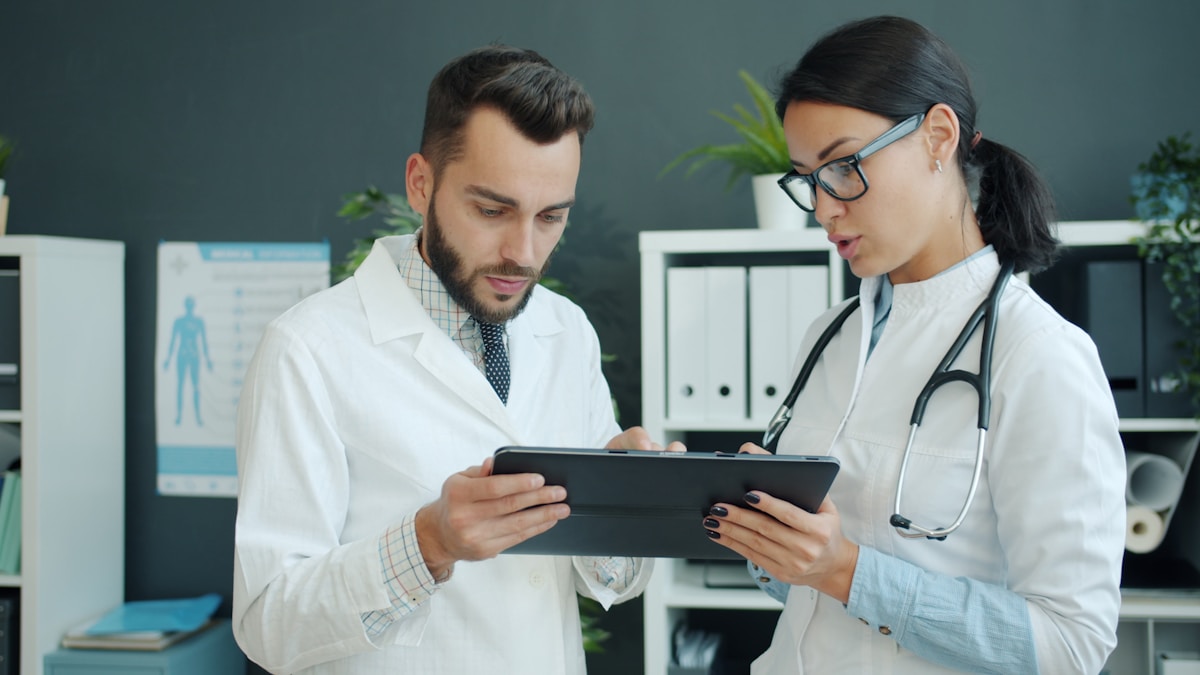 Medics in white gowns young man and woman are using tablet at work talking using gadget in office. Modern devices, pe...