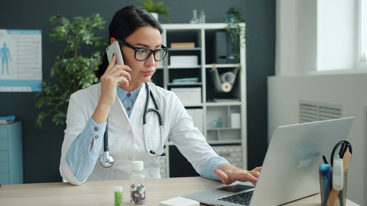 A clinician talking on a mobile phone while working on a laptop at a desk