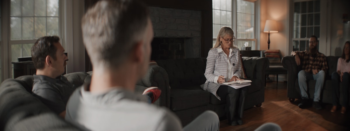 Wide view of a group seated together in a living room, a woman with glasses taking notes on a sofa while afternoon li...